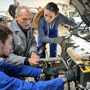 Auszubildende mit Ausbilder in einer Kfz-Werkstatt vor Auto mit geöffneter Motorhaube