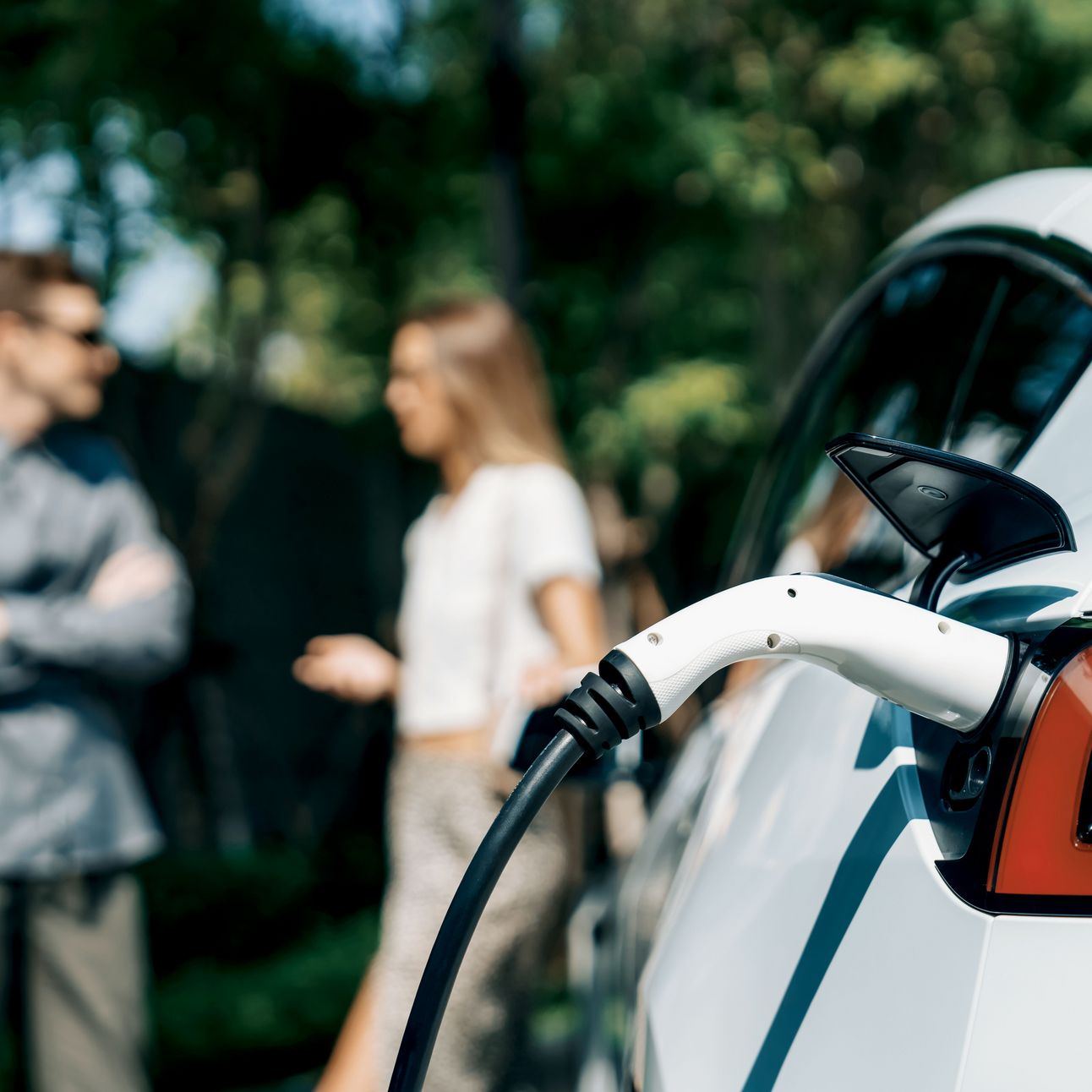 Auto hängt an der Ladesäule, ein Paar diskutiert im Hintergrund