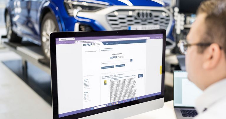 In moderner, lichtdurchfluteter Kfz-Werkstatt: Audi A8 e-tron auf der Hebebühne, Mitarbeiter recherchiert die passende Reparaturanleitung bei RepairPedia