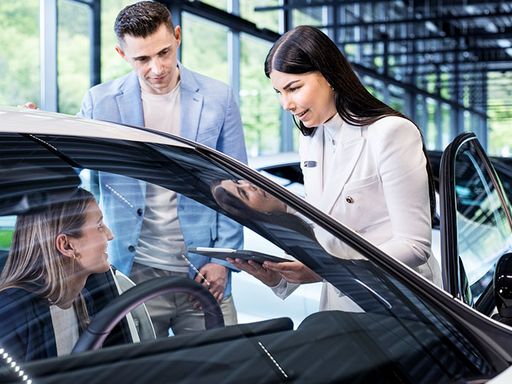 Autokauf in einem Autohaus-Showroom: Frau sitzt in Auto, Mann steht daneben und Verkäuferin gibt den beiden Infos zum Fahrzeug.