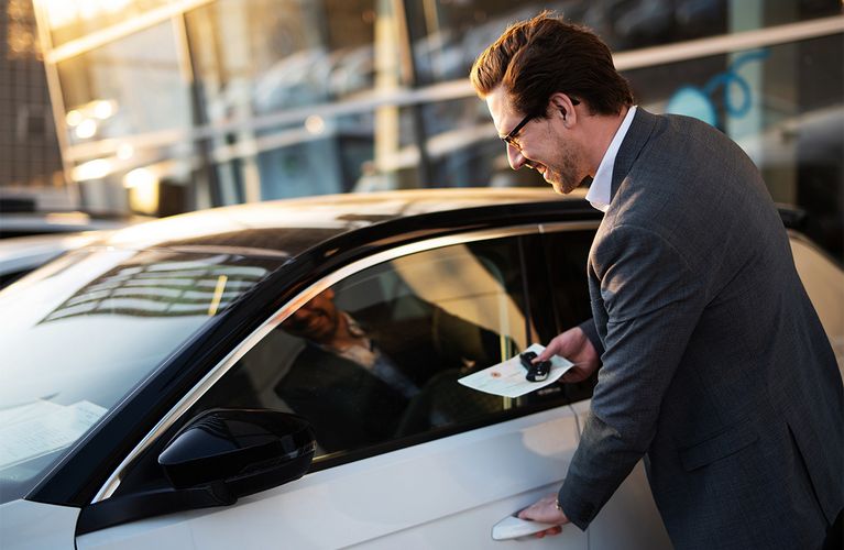 Ein Mann steht an einem Auto und hat den Schlüssel und Fahrzeugschein in der Hand. Er will gerade einsteigen und greift mit der anderen Hand zum Türgriff.