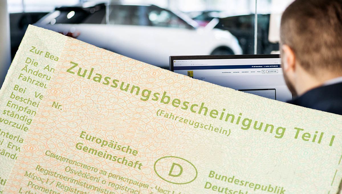Ansicht eines Fahrzeugscheins (Zulassungsbescheinigung Teil 1) im Vordergrund, dahinter sitzt der Mitarbeiter eines Autohauses an einem Computer