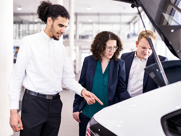 Autokauf: Ein Paar lässt sich im Autohaus von einem Verkäufer am Heck eines weißen Pkw beraten