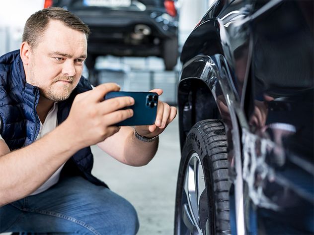 Ein Mann fotografiert mit seinem Handy den Unfallschaden (verkratzten Lack) an einem schwarzen Auto