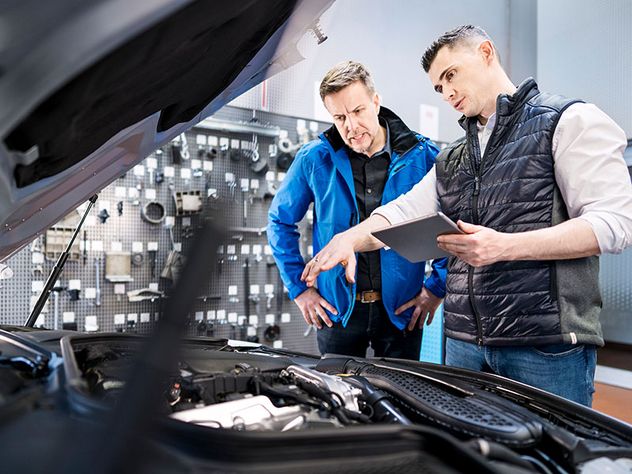 Szene in einer Kfz-Werkstatt: Automechaniker / Mechatroniker erklärt dem Kunden dem Kunden bei geöffneter Motorhaube den Reparaturbedarf an seinem Auto.