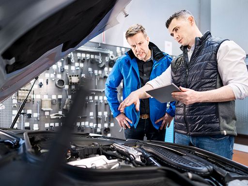 Szene in einer Kfz-Werkstatt: Automechaniker / Mechatroniker erklärt dem Kunden dem Kunden bei geöffneter Motorhaube den Reparaturbedarf an seinem Auto.