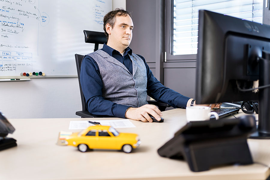 Mann im Büro vor Computer-Bildschirm, im Vordergrund ein gelbes Modell-Auto, im Hintergrund ein Whiteboard