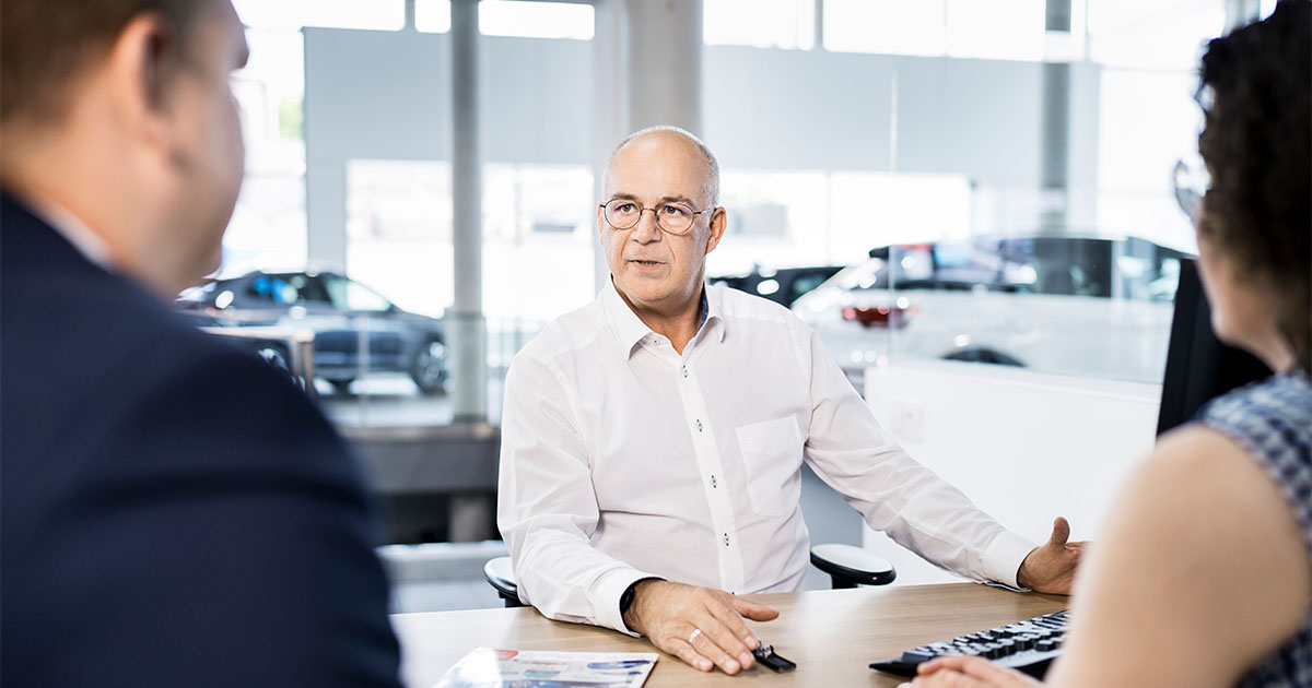 Drei Personen sitzen an einem Schreibtisch in einem Autohaus und unterhalten sich.