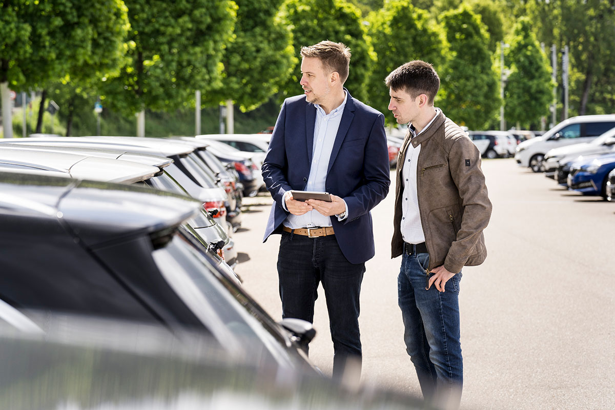 Neuwagen oder Gebrauchtwagen? Ein Auto-Käufer (= Kaufplaner) zusammen mit dem Mitarbeiter eines Autohandelsbetriebs schaut sich auf dem Hof abgestellte Fahrzeuge an.