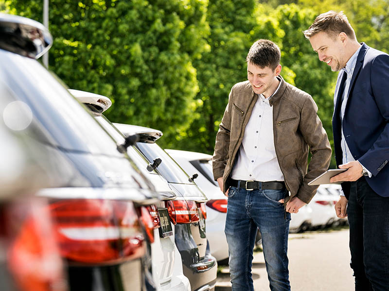 Ein Kaufinteressent lässt sich von Verkaufsmitarbeiter für Autokauf beraten. Beide stehen vor dem Heck von zum Verkauf angebotenen Pkw.