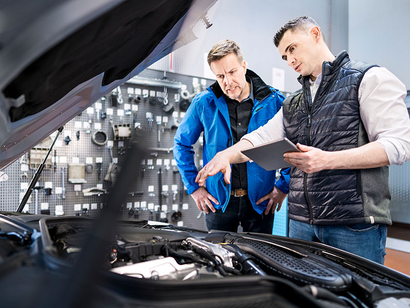 Szene in einer Kfz-Werkstatt: Automechaniker / Mechatroniker erklärt dem Kunden dem Kunden bei geöffneter Motorhaube den Reparaturbedarf an seinem Auto.
